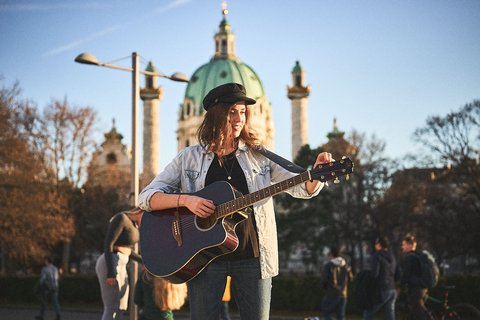 Eine junge Frau mit Gitarre steht vor einer historischen Architektur. Sie trägt eine Jacke und eine Mütze, während sie spielt und sich auf ihr Instrument konzentriert. Im Hintergrund sind Bäume und weitere Personen, die sich im Freien aufhalten, sichtbar.