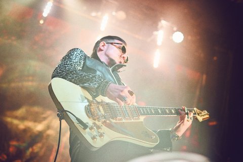 Ein Musiker spielt leidenschaftlich Gitarre auf einer Bühne. Er trägt eine Lederjacke mit Tiermuster und Sonnenbrillen. Im Hintergrund sind Lichter zu sehen, die eine lebhafte Atmosphäre erzeugen.