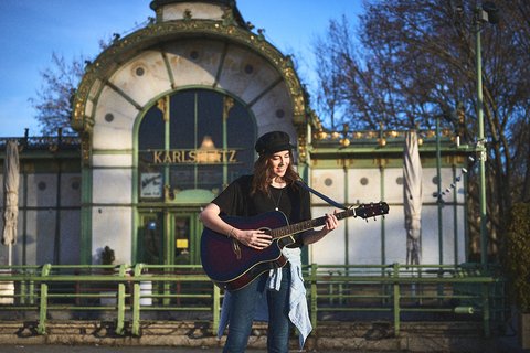 Eine junge Frau steht mit einer Gitarre in der Hand und singt. Im Hintergrund ist ein historisches Gebäude mit der Aufschrift „KARLSHAUS“ zu sehen. Der Himmel ist blau und die Szene wirkt sonnig und freundlich.