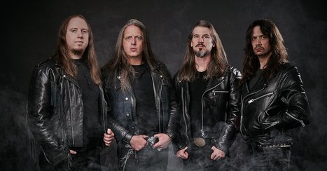 Vier Männer stehen nebeneinander, alle in schwarzen Lederjacken gekleidet. Sie schauen ernst in die Kamera. Der Hintergrund ist dunkel und leicht neblig, was eine rockige Atmosphäre schafft.