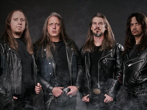 Vier Männer stehen nebeneinander, alle in schwarzen Lederjacken gekleidet. Sie schauen ernst in die Kamera. Der Hintergrund ist dunkel und leicht neblig, was eine rockige Atmosphäre schafft.