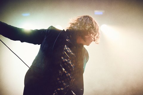 Ein Sänger auf der Bühne, der sich in die Musik vertieft. Sein Kopf ist zur Seite geneigt und die Haare fallen ihm ins Gesicht. Er trägt ein dunkelgrünes Jackett über einem gemusterten Hemd. Die Hintergrundbeleuchtung erzeugt eine stimmungsvolle Atmosphäre.