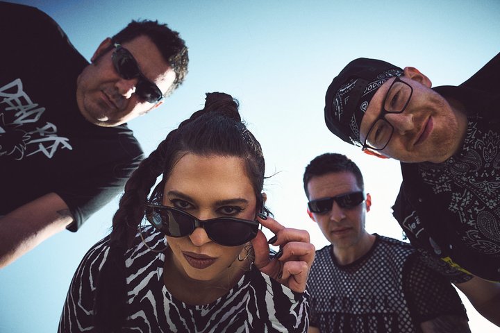 Vier Personen, cool posierend und von oben in die Kamera blickend, vor blauem Himmel.