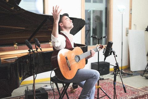 Ein Musiker sitzt auf einem Stuhl, spielt Gitarre und hebt eine Hand in die Luft. Hinter ihm ist ein Flügel sichtbar. Mikrofone stehen in seiner Nähe. Der Raum hat eine helle, kreative Atmosphäre.