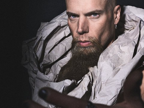 Ein maskuliner Mann mit Glatze und einem dichten Bart trägt einen auffälligen, zerknitterten Stoff um seinen Körper. Er schaut direkt in die Kamera mit einem intensiven Ausdruck und ausgestreckten Händen, die eine einladende Geste suggerieren. Der Hintergrund ist dunkel.