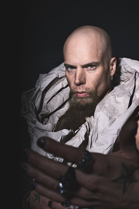 A bald man with a prominent beard is wearing a textured, layered garment. He gazes intensely at the viewer, extending his hands forward, adorned with multiple rings. The background is dark, enhancing the dramatic mood of the image.