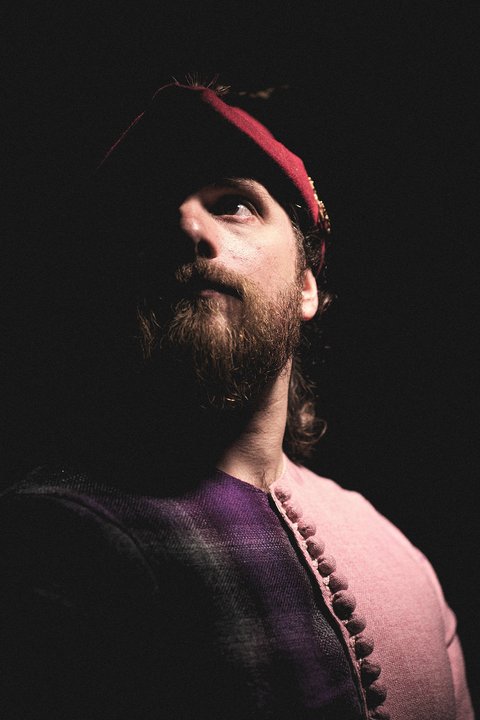 Ein Mann mit einem Bart trägt ein historisches Gewand in lila und rosa Farben. Er schaut nach oben, während ein schwaches Licht sein Gesicht von der Dunkelheit abhebt. Der Mann hat eine rote Kopfbedeckung auf.