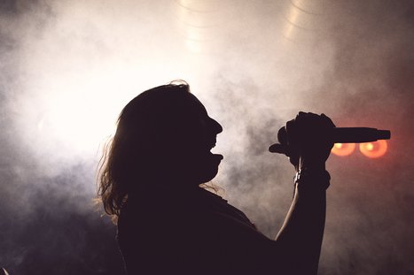 A silhouetted figure with long hair passionately sings into a microphone, surrounded by smoky mist and soft light. The atmosphere conveys energy and emotion, typical of a live music performance.