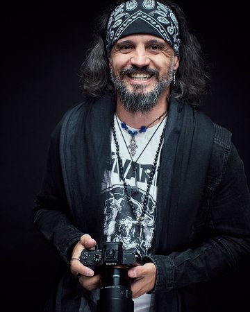 Ein lächelnder Mann mit einem Bandana und langen Haaren hält eine Kamera in der Hand. Er trägt ein schwarzes Oberteil und verschiedene Halsketten, vor einem neutralen, dunklen Hintergrund.