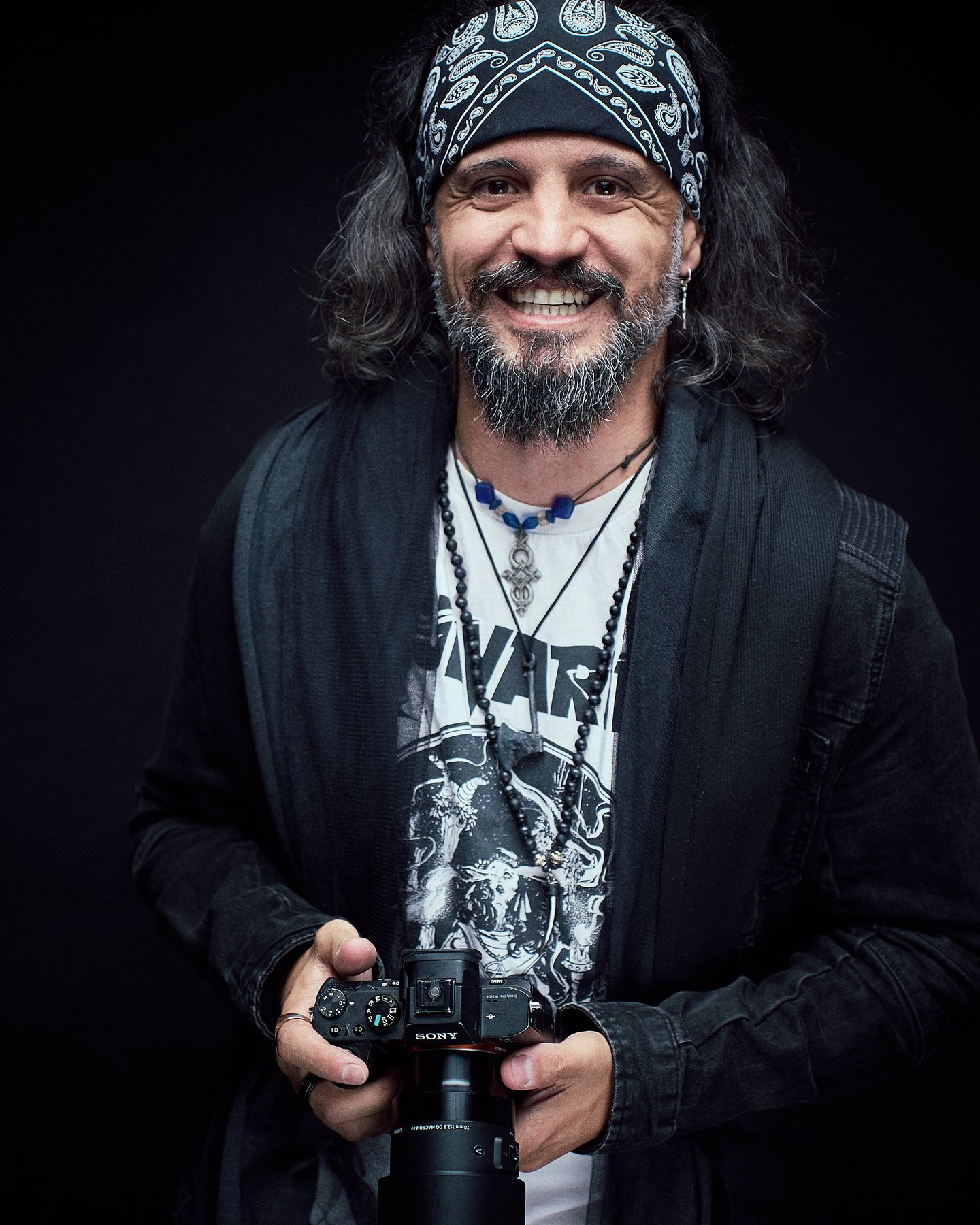 Ein lächelnder Mann mit einem Bandana und langen Haaren hält eine Kamera in der Hand. Er trägt ein schwarzes Oberteil und verschiedene Halsketten, vor einem neutralen, dunklen Hintergrund.