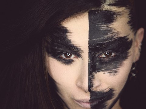 Eine Person mit halbem schwarz-weißen Gesichtsmalerei und langen, dunklen Haaren blickt direkt in die Kamera. Die rechte Gesichtshälfte ist in dunklen Farben gestaltet, während die linke Hälfte hell und unbemalt ist. Der Hintergrund ist schwarz.