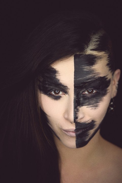 A close-up portrait of a person with half of their face painted black and the other half in a light color. The makeup features bold, sweeping strokes, creating a striking contrast against the person's dark hair. The background is black, emphasizing the dramatic facial art.