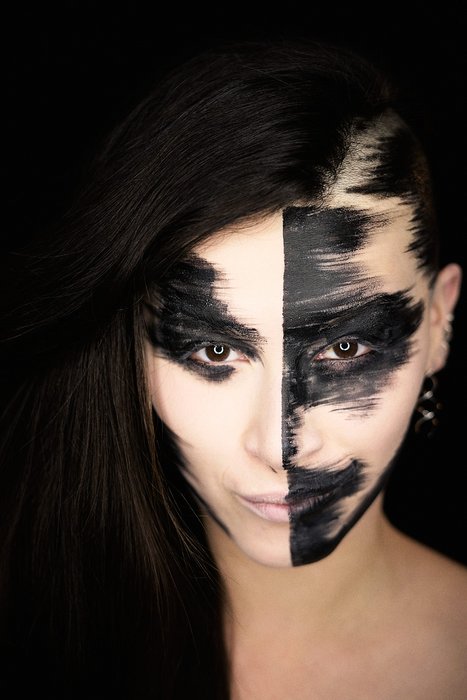 Arte Facial Abstrata em Preto e Branco Retrato de uma pessoa com maquiagem facial artística, metade do rosto pintada de branco e a outra metade de preto, criando um contraste marcante. A expressão é intensa e os olhos têm um olhar penetrante, enquanto o fundo é escuro, destacando a face.