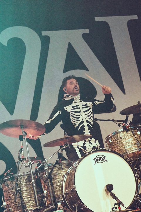 Ein Schlagzeuger in einem Skelettkostüm spielt leidenschaftlich Schlagzeug auf einer Bühne. Die Szene ist beleuchtet, und er konzentriert sich auf seine Performance, während er die Drums mit Energie schlägt.