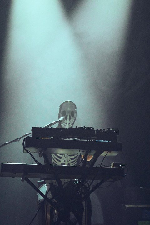 Ein Musiker steht hinter einem Keyboard und trägt ein Skelettkostüm. Der Raum ist schwach beleuchtet, mit Lichtstrahlen, die auf ihn fallen. Die Atmosphäre wirkt geheimnisvoll und eindringlich.