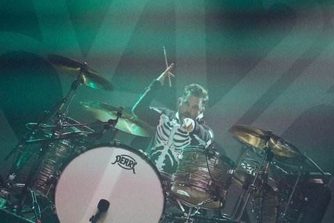 Ein Schlagzeuger in einem Skelettkostüm spielt auf seiner Trommel. Er hebt einen Drumstick und zieht die Aufmerksamkeit des Publikums auf sich. Die Atmosphäre ist energiegeladen und hell erleuchtet, passend zu einem Live-Konzert.