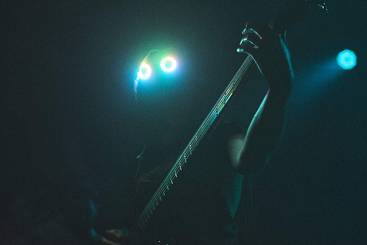 Ein Musiker spielt Bass auf einer dunklen Bühne, während gewellte Lichter seine Augen beleuchten. Die Silhouette des Künstlers ist erkennbar, aber das Gesicht ist im Schatten verborgen. Die Atmosphäre ist intensiv und mysteriös.