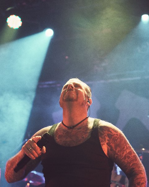 Ein tätowierter Sänger trägt ein schwarzes Tanktop und steht auf einer Bühne, während er die Arme leicht hebt und den Kopf zur Decke neigt. Bunte Lichter strahlen auf ihn, während eine rockige Atmosphäre herrscht.