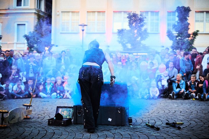 Eine Person führt eine Darbietung auf einem Platz durch, umgeben von einer großen Menge von Zuschauern. Es gibt Rauch und Requisiten auf dem Boden. Die Zuschauer schauen gespannt zu, während die Künstlerin auf einen Koffer blickt.