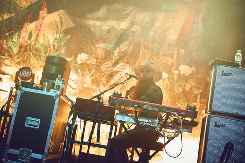 Ein Musiker sitzt an einem Keyboard auf einer Bühne, umgeben von Lichtern und einem farbenfrohen Hintergrund. Er trägt einen Hut und hat einen langen Bart. Im Vordergrund sind Musikequipment und Lautsprecher zu sehen. Die Atmosphäre wirkt lebhaft und energiegeladen.