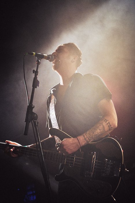 A musician stands on stage, singing into a microphone while playing an electric guitar. The scene is illuminated by soft, dramatic lighting, creating a lively atmosphere. The artist has visible tattoos on their arm and appears focused on their performance.