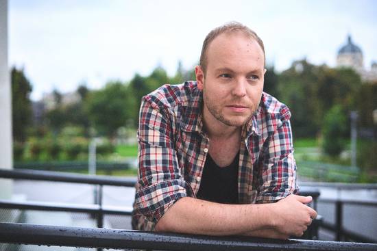 Ein Mann mit kurzen Haaren trägt ein kariertes Hemd und lehnt an einem Geländer. Er schaut nachdenklich, während er in eine grüne Landschaft mit Bäumen und einem Gebäudekomplex im Hintergrund blickt. Die Szene vermittelt eine ruhige, nachdenkliche Stimmung.