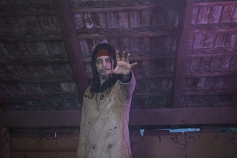Eine blutige, geisterhafte Figur steht in einer baufälligen Umgebung und streckt eine Hand aus. Der Ausdruck ist eindringlich und unheimlich, mit zerzausten Haaren und einem schmutzigen Gewand. Der Hintergrund ist schummrig, was die unheimliche Atmosphäre verstärkt.