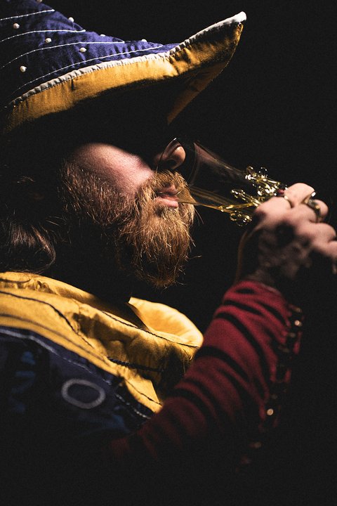 Ein Mann mit einem bunten Hut und einem langen Bart hält ein Glas in der Hand. Er hat ein nachdenkliches Gesicht und scheint das Glas zu betrachten. Der Hintergrund ist dunkel, was den Fokus auf seine Gesichtszüge und das Glas lenkt.
