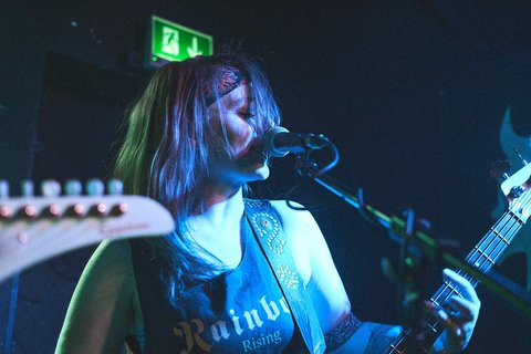 Eine Musikerin mit längeren Haaren steht vor einem Mikrofon und spielt Bass. Sie trägt ein schwarzes Shirt mit dem Aufdruck "Rainbow" und hat einen Haarreifen. Die Bühne ist schwach beleuchtet, was eine lebendige Atmosphäre schafft.
