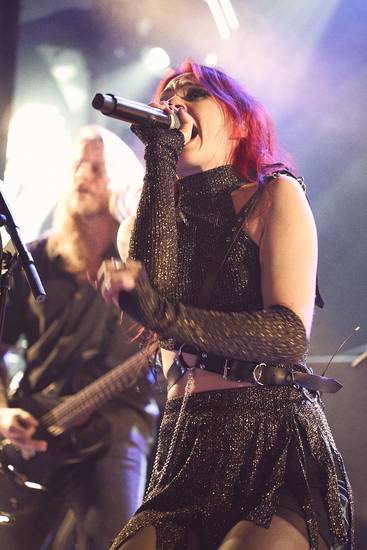 Eine Sängerin mit roten Haaren performt leidenschaftlich auf der Bühne, während ein Musiker mit einer Gitarre im Hintergrund spielt. Die Atmosphäre ist lebhaft und energiegeladen. Die Sängerin trägt ein glitzerndes Outfit und nutzt ein Mikrofon.