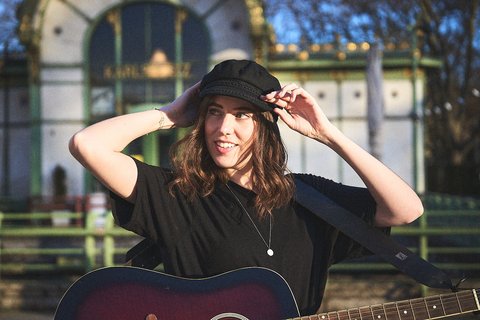 Eine junge Frau mit schulterlangen, lockigen Haaren trägt ein schwarzes T-Shirt und eine Mütze. Sie hält eine Gitarre in der linken Hand und lächelt, während sie sich an die Mütze fasst. Im Hintergrund sind Anzeichen eines Parks oder eines offenen Geländes sichtbar.