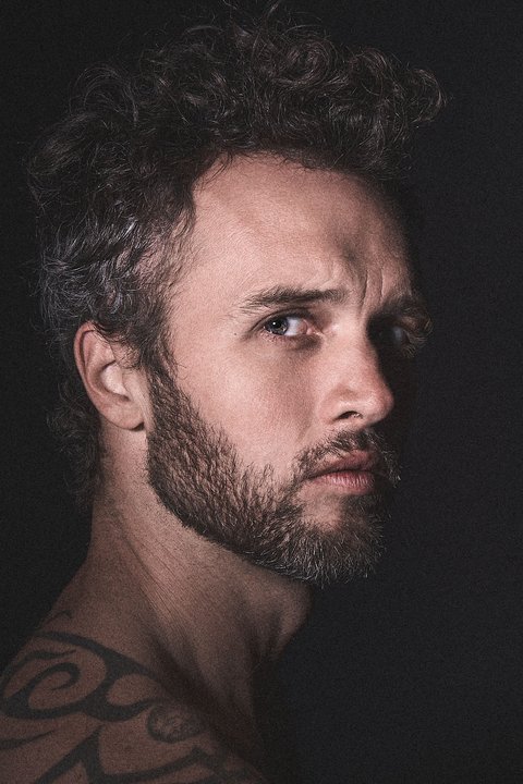 A man with a beard and tattoos looks seriously at the camera.