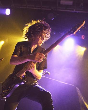 Ein Musiker mit lockigem Haar spielt leidenschaftlich eine E-Gitarre auf einer Bühnen mit bunten Lichtern. Er trägt ein schwarzes, glitzerndes Hemd und beugt sich leicht vor, während er sich auf seinePerformance konzentriert. Die Stimmung der Aufführung ist energiegeladen.