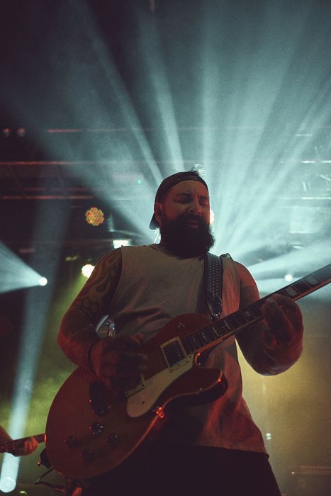 A musician with a beard plays an electric guitar on stage. Bright beams of light radiate behind him, creating a dynamic atmosphere. The performer appears focused and engaged, contributing to the lively ambiance of a concert.