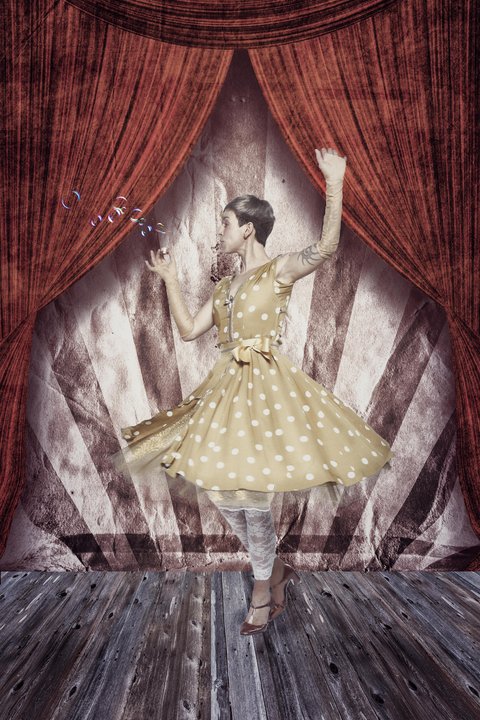 Eine tanzende Frau in einem gelben, gepunkteten Kleid steht auf der Bühne vor rotem Vorhang. Sie hebt einen Arm und hält ein Seifenblasenbehälter in der anderen Hand. Die Szenerie vermittelt ein Gefühl von Freude und Verspieltheit.