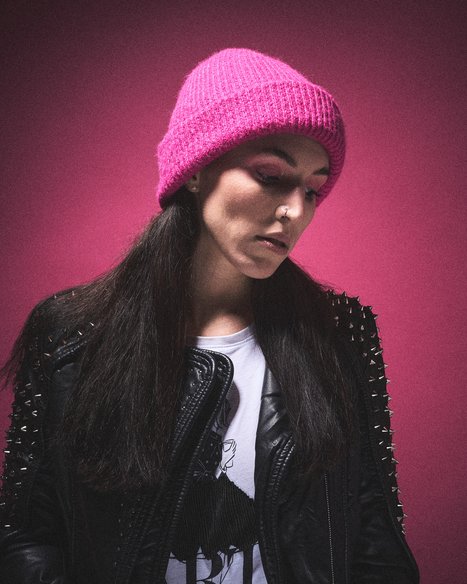 Edgy Chic: Woman in Pink Beanie and Spiked Leather Jacket A young woman with long dark hair wears a bright pink beanie and a black leather jacket adorned with studs. She looks down thoughtfully against a vivid pink background, creating a striking and stylish contrast.