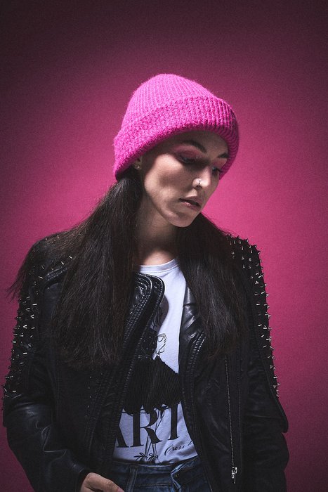 Eine junge Frau mit langen, dunklen Haaren trägt eine pinke Mütze und eine schwarze Lederjacke mit Nieten. Sie schaut nach unten, vor einem einfarbigen, pinken Hintergrund. Ihr Ausdruck vermittelt Nachdenklichkeit oder Melancholie.