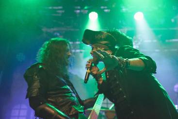 Zwei Musiker stehen auf der Bühne, umgeben von grünen Lichtstrahlen. Einer der Künstler trägt ein Piratenkostüm und hält ein Mikrofon, während er mit dem anderen spricht. Beide haben eine leidenschaftliche Bühnenpräsenz.