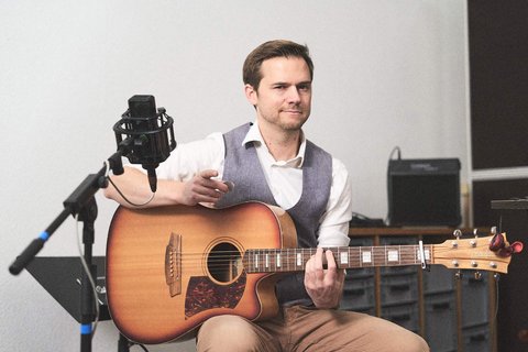 Ein Mann mit einem freundlichen Gesicht sitzt mit einer Akustikgitarre in der Hand. Er trägt ein weißes Hemd und eine graue Weste. Im Hintergrund sind Mikrofon und einige Möbelstücke sichtbar, während er spielt und anspricht.