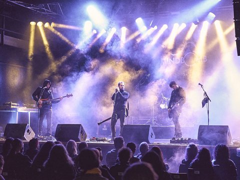 Eine Band tritt auf einer Bühne auf, umgeben von bunten Lichtern und Nebel. Der Sänger steht im Mittelpunkt und wird von zwei Instrumentalisten flankiert. Das Publikum sitzt vor der Bühne und schaut gebannt zu.