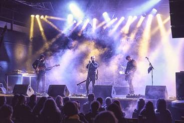 Eine Band tritt auf einer Bühne auf, umgeben von bunten Lichtern und Nebel. Der Sänger steht im Mittelpunkt und wird von zwei Instrumentalisten flankiert. Das Publikum sitzt vor der Bühne und schaut gebannt zu.