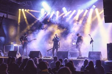 Eine Band tritt auf einer Bühne auf, umgeben von bunten Lichtern und Nebel. Der Sänger steht im Mittelpunkt und wird von zwei Instrumentalisten flankiert. Das Publikum sitzt vor der Bühne und schaut gebannt zu.