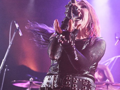 Eine Sängerin auf der Bühne mit einem Mikrofon in der Hand, trägt schwarze Kleidung mit Nieten und Ketten. Sie hat lange, pinke Haare und wirkt leidenschaftlich beim Singen. Im Hintergrund sind verschwommene Instrumente zu erkennen, die auf eine Rock- oder Metal-Performance hindeuten.