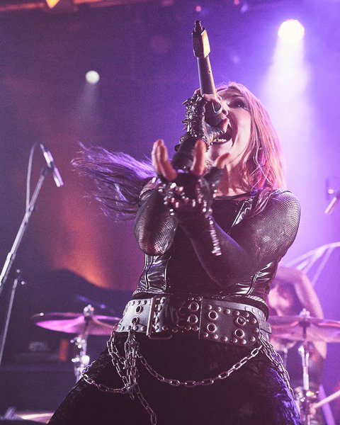 A person with long, flowing hair performs passionately on stage, holding a microphone. They are dressed in a black outfit adorned with chains and accessories, surrounded by colorful stage lights. The energetic atmosphere reflects their powerful vocal performance.