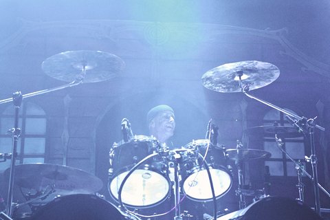 Ein Schlagzeuger spielt in einem schwach beleuchteten Raum. Er trägt einen Hut und konzentriert sich auf sein Spiel. Im Hintergrund sind verschwommene Gegenstände zu erkennen, die zur Bühnenatmosphäre beitragen.