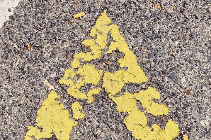 Ein gelbes, abgenutztes Pfeilsymbol auf grauem Asphalt. Die Farbe ist stellenweise abgeblättert, was dem Symbol ein alterndes Aussehen verleiht.
