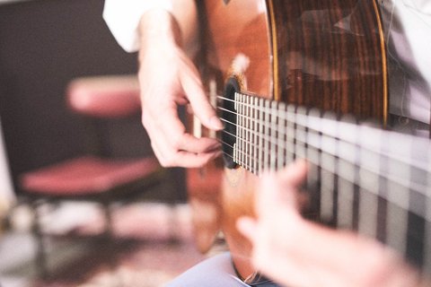 Eine Hand spielt auf den Saiten einer akustischen Gitarre, während eine andere Hand den Hals der Gitarre greift. Im Hintergrund ist ein unscharfer Stuhl sichtbar, was eine gemütliche musikalische Atmosphäre vermittelt.
