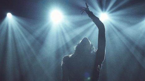 A silhouetted figure stands in front of bright stage lights, reaching upward with one arm. The scene captures a moment of artistic expression and performance, creating a dramatic and energetic atmosphere.