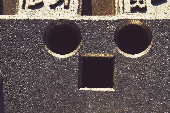 Der Inhalt zeigt einen Block mit zwei runden Löchern und einem quadratischen Loch darunter, die zusammen ein Gesicht mit Augen und Mund bilden. Die Oberfläche des Blocks ist strukturiert und schwarz, was dem Gesicht einen markanten Ausdruck verleiht.