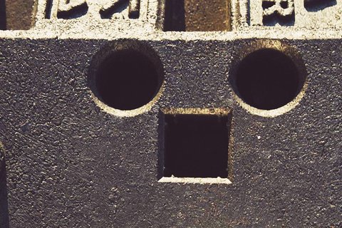 Der Inhalt zeigt einen Block mit zwei runden Löchern und einem quadratischen Loch darunter, die zusammen ein Gesicht mit Augen und Mund bilden. Die Oberfläche des Blocks ist strukturiert und schwarz, was dem Gesicht einen markanten Ausdruck verleiht.